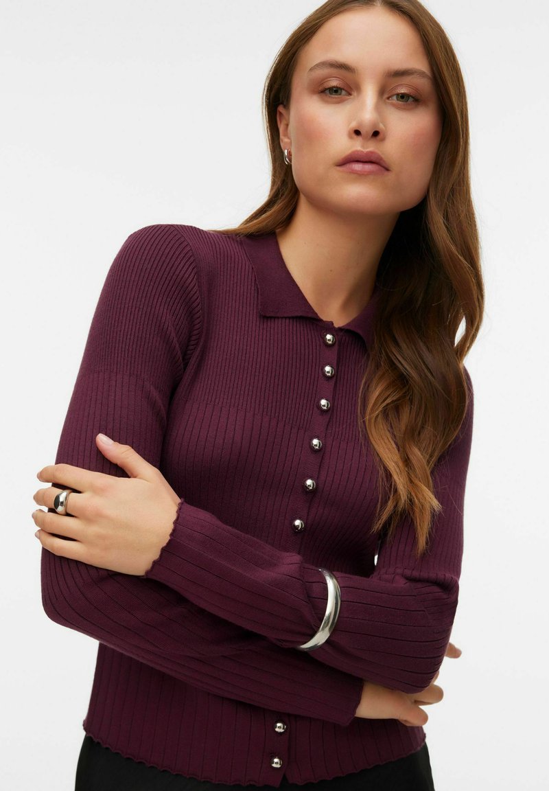 Bordo spalvos kotuotas megztinis su apykakle ir sidabro spalvos sagų užsegimais. Ilgos rankovės su banguotais kraštais. Lygus tekstūra.