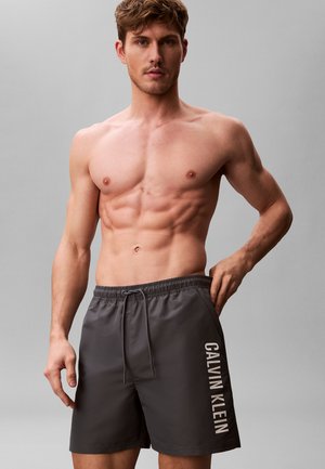 Muskularny, nagi do pasa mężczyzna w ciemnoszarych kąpielówkach Calvin Klein, stojący na jednolitym szarym tle, z ręką spoczywającą przy kieszeni.