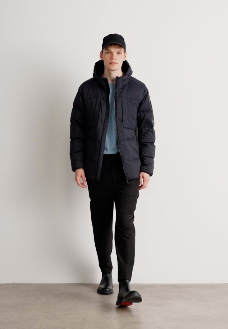 Doudoune noire avec fermeture éclair et deux grandes poches, associée à une chemise bleu clair et un pantalon noir. Une casquette et des chaussures noires complètent le look.