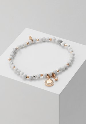 Bracelet de perles marbrées blanches avec de petites perles dorées et deux pendentifs en or, l'un rond avec des cristaux, présenté sur un cube blanc.