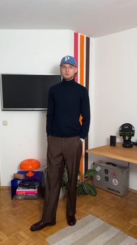 Mornarica pletena turtleneck, rjave hlače in burgundske čevlje. Modra kapa z logotipom. Minimalistična dekoracija vključuje oranžno svetilko in sobne rastline.