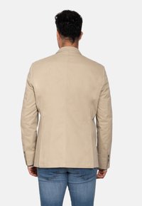Blazer entallado beige con tela suave, abertura sencilla y puños abotonados. Llevado sobre jeans azules, visto desde atrás.