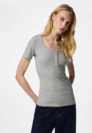 Ξανθιά γυναίκα που φοράει ανοιχτό γκρι ριμπ κοντομάνικο τοπ με κουμπιά και σκούρο μπλε τζιν, στέκεται με τα χέρια πίσω από την πλάτη.