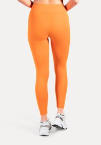 Orange leggings med hög midja, slät textur och hel längd. Bärs med vita sportskor och visar en elegant design.