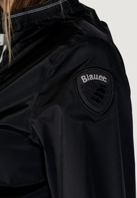 Giacca impermeabile nera con colletto alto, polsini elastici e una patch con il logo sulla manica. Texture liscia con accenti riflettenti.