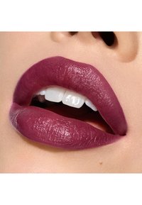 Rouge à lèvres mat de couleur baies foncées sur des lèvres charnues, avec une texture lisse et un éclat subtil, mettant en valeur des dents blanches et un teint clair en arrière-plan.
