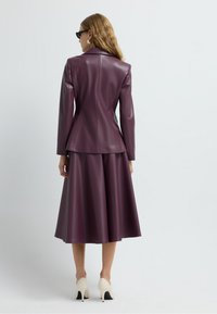 Blazer en simili cuir bordeaux avec une silhouette ajustée, associé à une jupe assortie jusqu'aux genoux et des chaussures à talons hauts blanches.