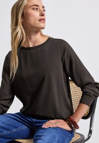 Lässig geschnittene dunkelbraune Bluse mit Rundhalsausschnitt und langen Ärmeln. Glatte Textur, kombiniert mit blauen Jeans und gestapelten Armbändern.