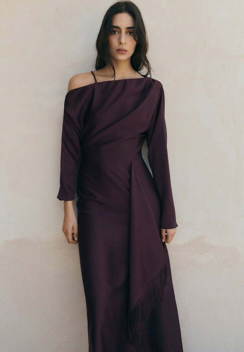 Giovane donna in piedi contro un muro beige, indossa un lungo abito viola scuro con una spalla scoperta e dettagli con frange sulla gonna.