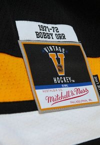 Schwarzes und gelbes Hockeytrikot-Tag mit "1971-72 BOBBY ORR", sowie den Logos "VINTAGE HOCKEY" und "Mitchell & Ness" in Rot und Blau.