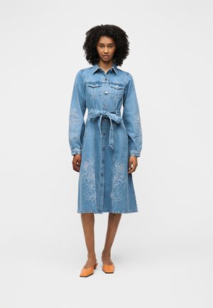 Une femme se tient debout portant une robe en jean mi-longue avec un devant à boutons, une taille ceinturée, une broderie florale, des manches longues et des chaussures orange à bout pointu.