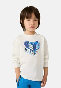 T-shirt bianco a maniche lunghe con un design grafico di testo colorato, caratterizzato da blocchi blu e faccine sorridenti, abbinato a pantaloni blu.