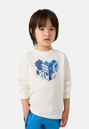 T-shirt bianco a maniche lunghe con un design grafico di testo colorato, caratterizzato da blocchi blu e faccine sorridenti, abbinato a pantaloni blu.