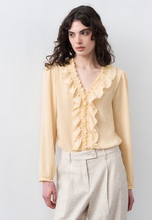 Γυναίκα που φοράει μια ανοιχτό κίτρινη μπλούζα με μακριά μανίκια και βολάν στο V-λαιμό και ανοιχτό μπεζ ψηλόμεσα παντελόνια, στέκεται μπροστά σε απλό φόντο.