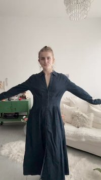 Žena s plavom kosom u dugoj tamnoj traper haljini stoji raširenih ruku u svijetloj dnevnoj sobi s bijelom sofom i zelenim ormarom.