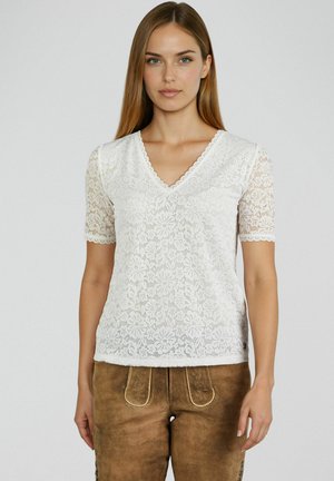 Vrouw die een wit topje met bloemenkant en korte mouwen draagt, met een geschulpte V-hals en bruine suède broek, rechtstaand tegen een effen lichte achtergrond.