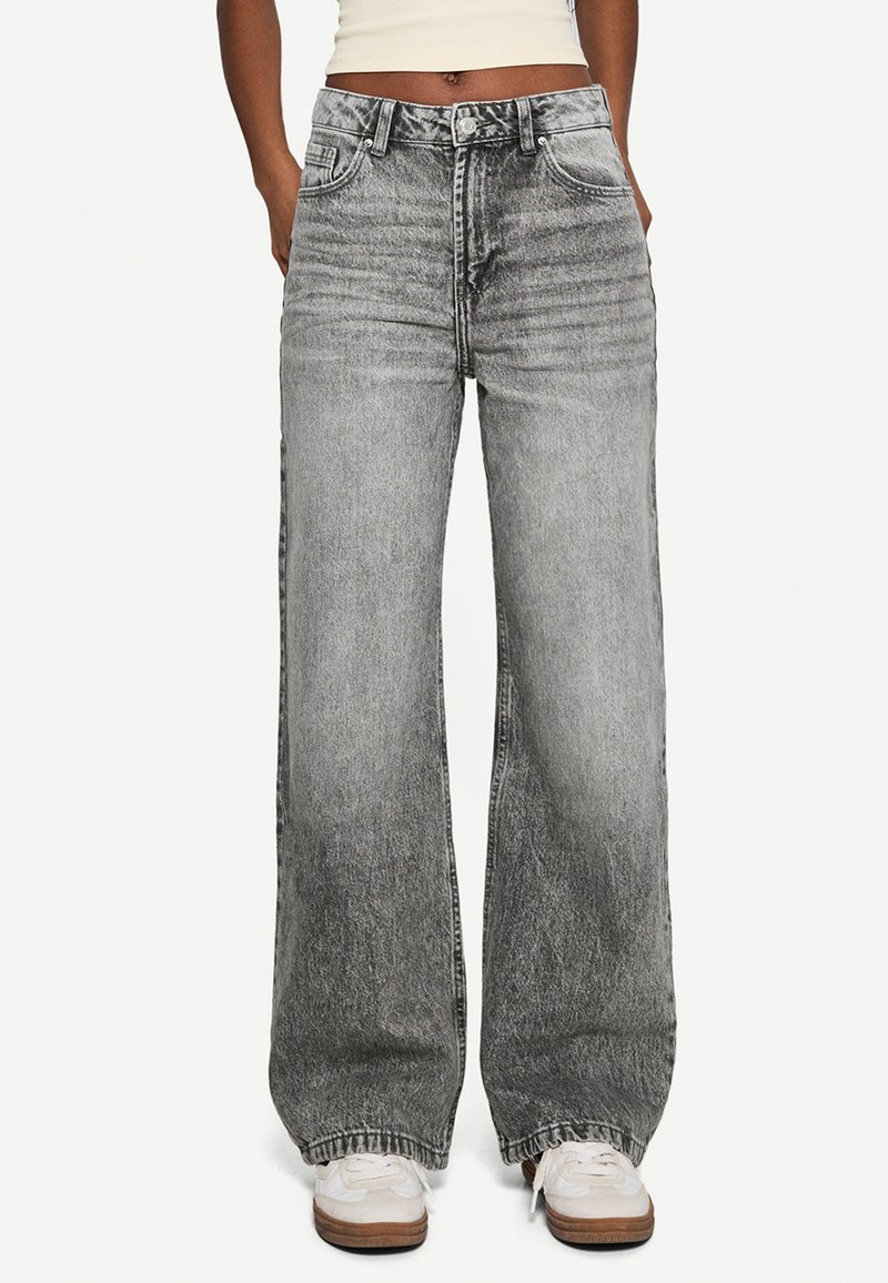 Weitbeinige Jeans aus verblichenem grauem Denim, mit hoher Taille, frontalen Taschen und subtilen Used-Effekten. Kombiniert mit weißen Sneakers.