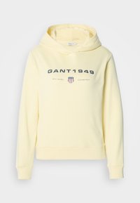 Žltá mikina s kapucňou z jemnej látky s potlačeným logom "GANT 1949" a motívom štítu na hrudi. Dlhé rukávy, rebrované manžety.