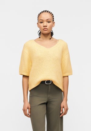 Donna con capelli intrecciati che indossa un maglione lavorato a maglia giallo morbido a maniche corte, infilato leggermente in pantaloni oliva a vita alta con cintura nera.