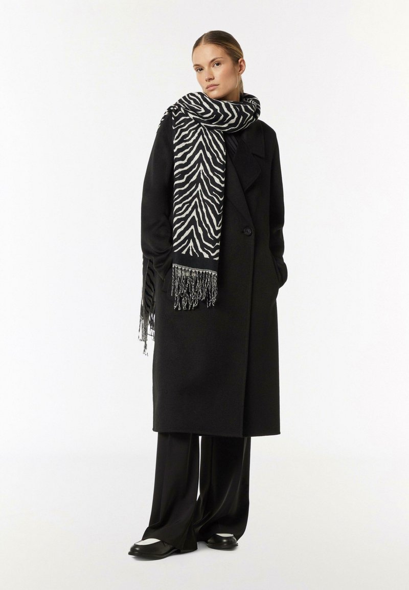 Black overcoat with large lapels, paired with a zebra-patterned black and white scarf featuring fringed ends. Loose-fitting black trousers and footwear.
