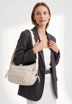 Vrouw die een donker colbert draagt over een witte jurk met een gevlochten riem, met een beige schoudertas met het "GWV"-logo.