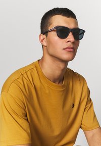 Ochelari de soare negri cu rame pătrate, având o dâra subtilă roșie și albastră pe brate. Purtați cu un tricou galben muștar cu mânecă scurtă.