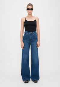 Svart ärmlös topp, midjehög vida blå jeans, textilmaterial och svarta platta skor med vita accenter. Minimalistisk design.