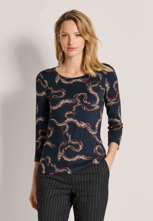 Blonde vrouw draagt een marineblauwe top met lange mouwen met een slangenpatroon en donkere broek met fijne strepen, staand met één hand in de zak.