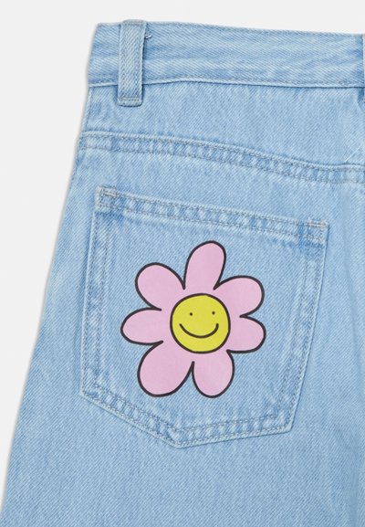 Denim azul claro com um patch de flor rosa e um rosto sorridente amarelo no bolso de trás. Textura suave e corte clássico.