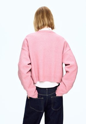 Gekürzter rosa Pullover mit geripptem Saum und weiten Ärmeln, getragen über einem weißen Hemd, kombiniert mit dunkelblauen Jeans. Sanfte Textur, minimales Design.