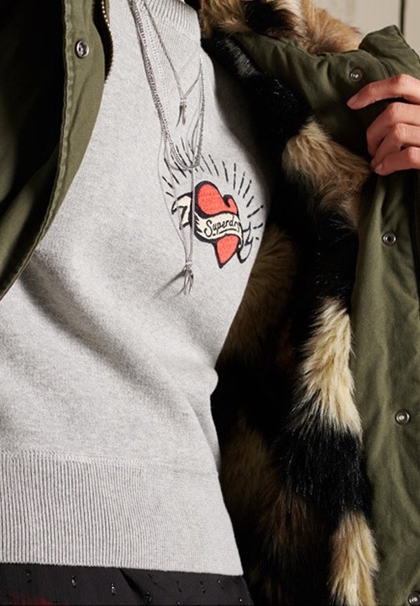 Maglione grigio con un cuore rosso e il design del testo "Superdry", indossato sotto un giubbotto verde con cappuccio foderato in pelliccia e collane d'argento.