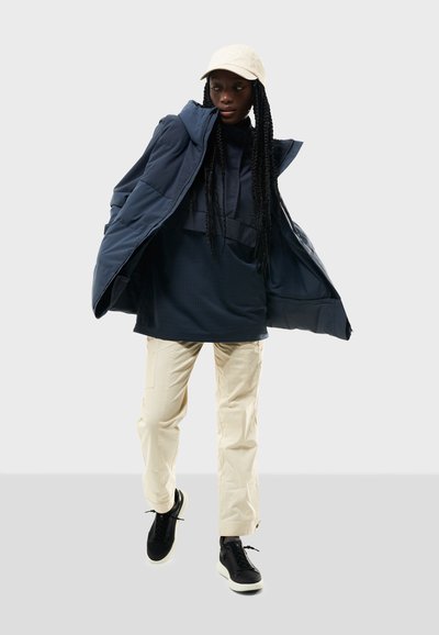 Marinblå vadderad jacka, svart långärmad tröja, beige cargobyxor och svarta sneakers. Beigekepsen fulländar outfiten. Släta texturer och funktionell design.