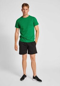 Grön bomullströja med korta ärmar och rund halsringning, ihop med svarta shorts, står framför en enkel bakgrund, bärande svarta träningsskor.