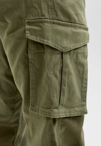 Primo piano della tasca di pantaloni cargo verde oliva con patta e cuciture visibili su tessuto caratterizzato da lievi pieghe.
