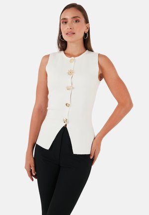 White sleeveless top with a textured finish, featuring five gold decorative buttons and a split hem design. Paired with black pants.