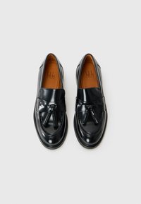 Mocassini in pelle verniciata nera con un accento a nappine, punta rotonda e soletta marrone a contrasto. Caratterizzati da una suola texture e un design elegante.