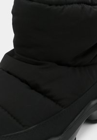 Fekete szigetelt csizma, amely párnázott mintával, sima anyaggal, lekerekített felsőrésszel és strapabíró gumitalppal rendelkezik a tapadás érdekében.