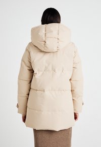 Veste en duvet beige avec une grande capuche, motif matelassé et ourlet cintré. Matériau rigide avec une texture lisse, dotée de manches longues.
