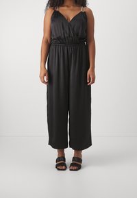 Mono negro de satén con diseño envolvente, tirantes ajustables, cintura elástica y piernas anchas recortadas, combinado con sandalias negras con tiras.