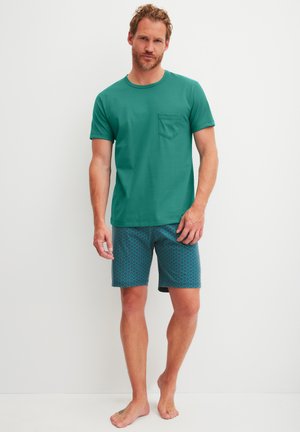 Mann steht barfuß und trägt ein türkisfarbenes kurzärmeliges Hemd mit Brusttasche sowie gemusterte türkisfarbene Shorts vor weißem Hintergrund.