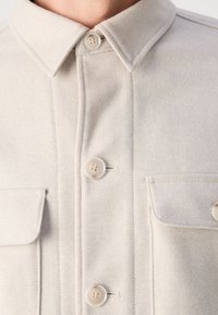 Camisa de lana en tono beige claro con collar puntiagudo, que cuenta con tres botones, dos bolsillos en el pecho y detalles de costura visibles. Textura suave.