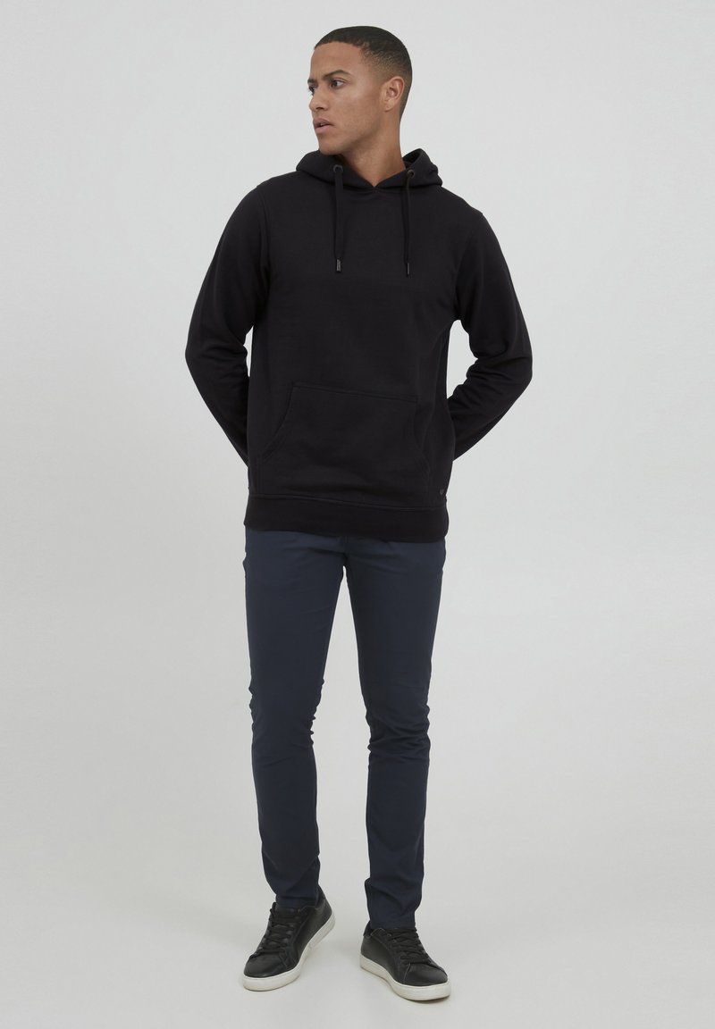 Felpa nera in cotone con tasca frontale e cappuccio con cordino, abbinata a pantaloni slim-fit blu navy e sneakers nere con suola bianca.