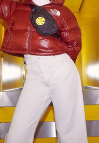 Czerwona kurtka puchowa z wyraźnym przeszyciem i logo na lewej piersi, połączona z jasnobeżowymi spodniami o wysokim stanie oraz czarną saszetką.