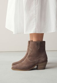 Brune ruskindsankelstøvler med et buet design, spids snude og stablet træsålshæl. Detaljeret syning og en lille rem ved anklen.