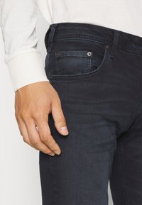 Jeans de mezclilla azul oscuro con una vista en primer plano de una mano descansando en el muslo, mostrando detalles del botón y el bolsillo.