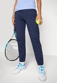 Person som har på seg marineblå joggebukser og hvite sneakers, holder en tenniskølle og en grønn tennisball foran en ensfarget bakgrunn.