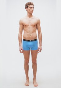 Νεαρός άνδρας με σγουρά μαλλιά που φοράει ανοιχτό μπλε εσώρουχα Polo Ralph Lauren, όρθιος ξυπόλητος μπροστά σε λευκό φόντο.