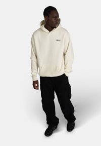 Felpa con cappuccio crema e tasca frontale, con la scritta "CRYSTAL" in testo nero. Abbinata a pantaloni cargo neri e calzature nere.