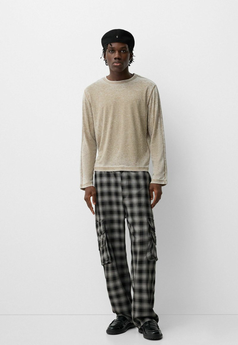 Camisa de manga larga beige con un acabado texturizado, combinada con pantalones cargo de cuadros en negro y gris, que cuentan con bolsillos laterales y un corte relajado. Se llevan zapatos negros.