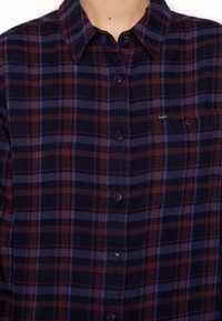 Camicia di flanella a quadri in blu navale, rosso e viola. Presenta un colletto con bottoni, chiusura con bottoni e una piccola tasca sul petto con logo.